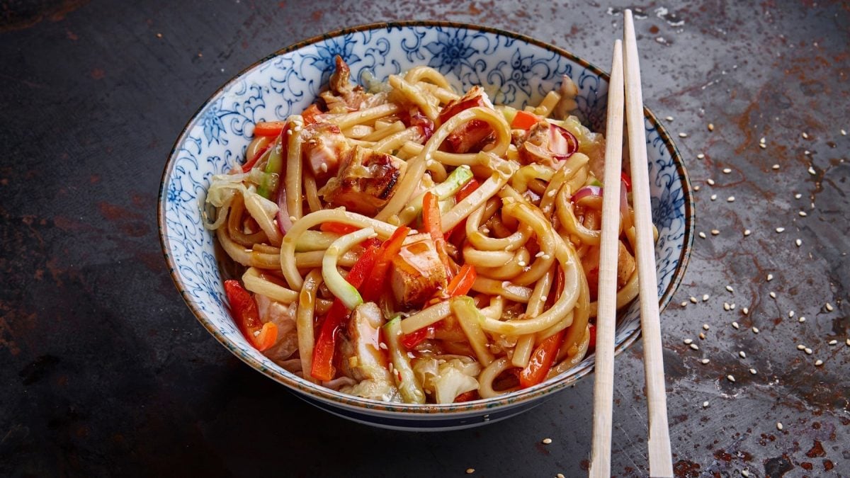 Eine weiß-blaue Schüssel auf dunklem Untergrund mit den Udon-Nudeln und zwei Essstäbchen auf der rechten Seite der Schüssel.
