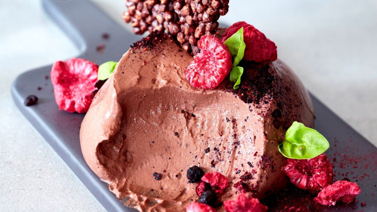 Eine Portion vegane Schokomousse mit getrockneten Himbeeren und Krokant und Basilikum auf einem Brettchen, heller Hintergrund.