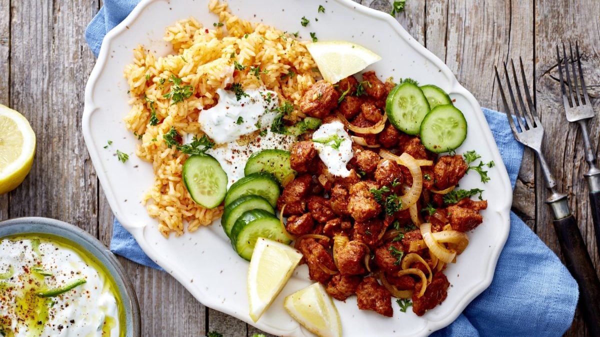 Eine weiße Platte mit vegetarischem Gyros, Reis, Zitronen und Gurken auf einem blauen Geschirrtuch auf einem hölzernen Hintergrund. Links unten ein Joghurt-Dip, rechts zwei Gabeln.