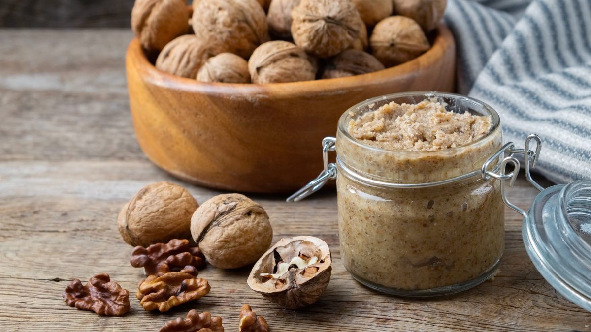 Frontal: Auf einem Untergrund aus Holz steht ein Glas Walnuss Pesto. Drum herum liegen ganze und aufgebrochene Walnüsse.