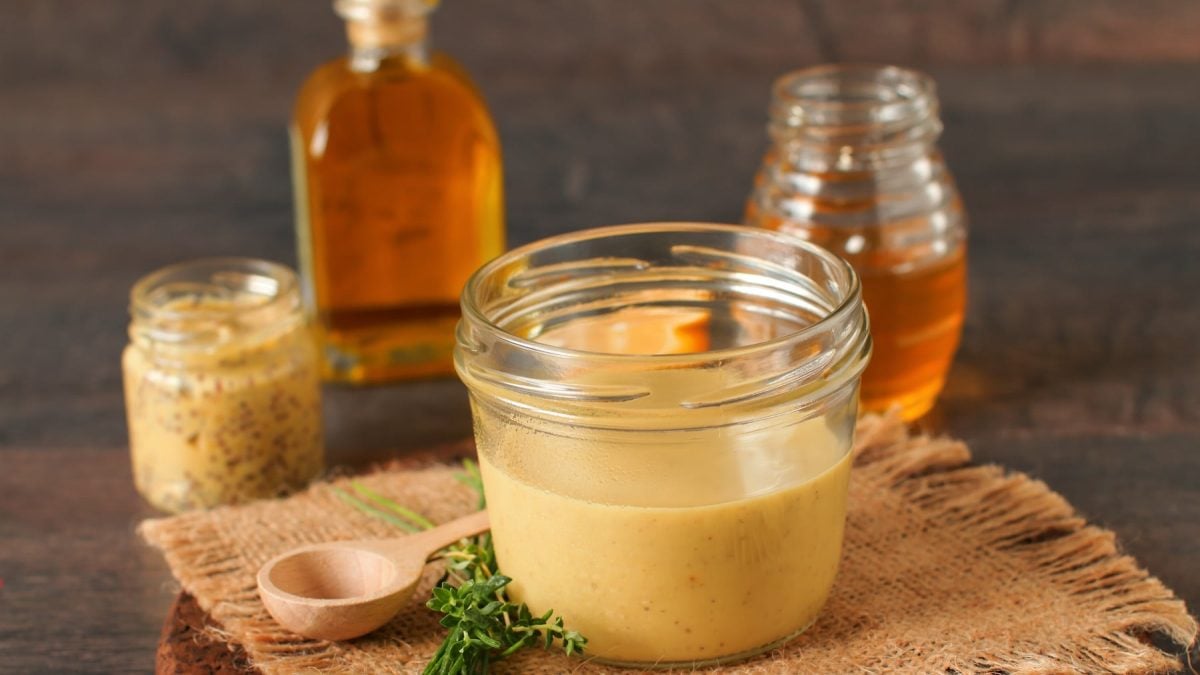 Ein Weckglas mit der Whiskey-Senf-Sahnesauce auf einem Holztisch mit einem Glas Honig, einem Glas grobem Senf und einer Flasche Whiskey.