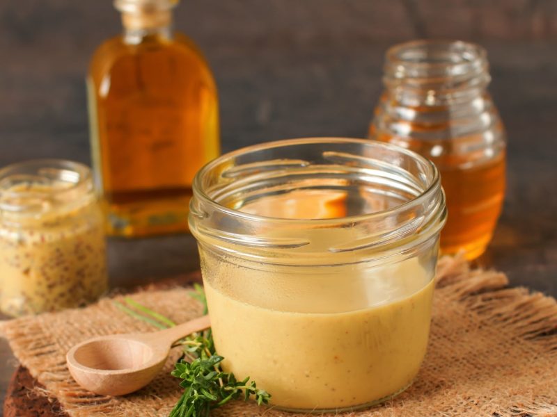 Ein Weckglas mit der Whiskey-Senf-Sahnesauce auf einem Holztisch mit einem Glas Honig, einem Glas grobem Senf und einer Flasche Whiskey.