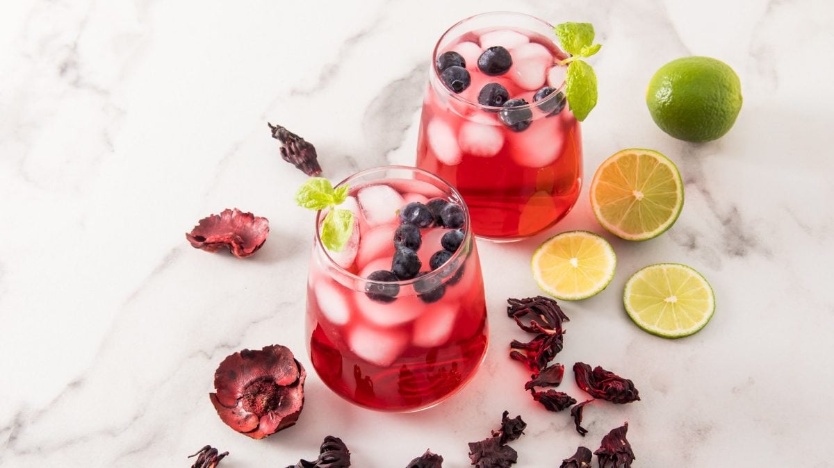 Zwei Gläser gefüllt mit dem Hibiskus-Eistee Agua de Jamaica, Eiswürfeln und Blaubeeren, drumherum getrocknete Hibiskusblüten, Limetten und Blaubeeren.