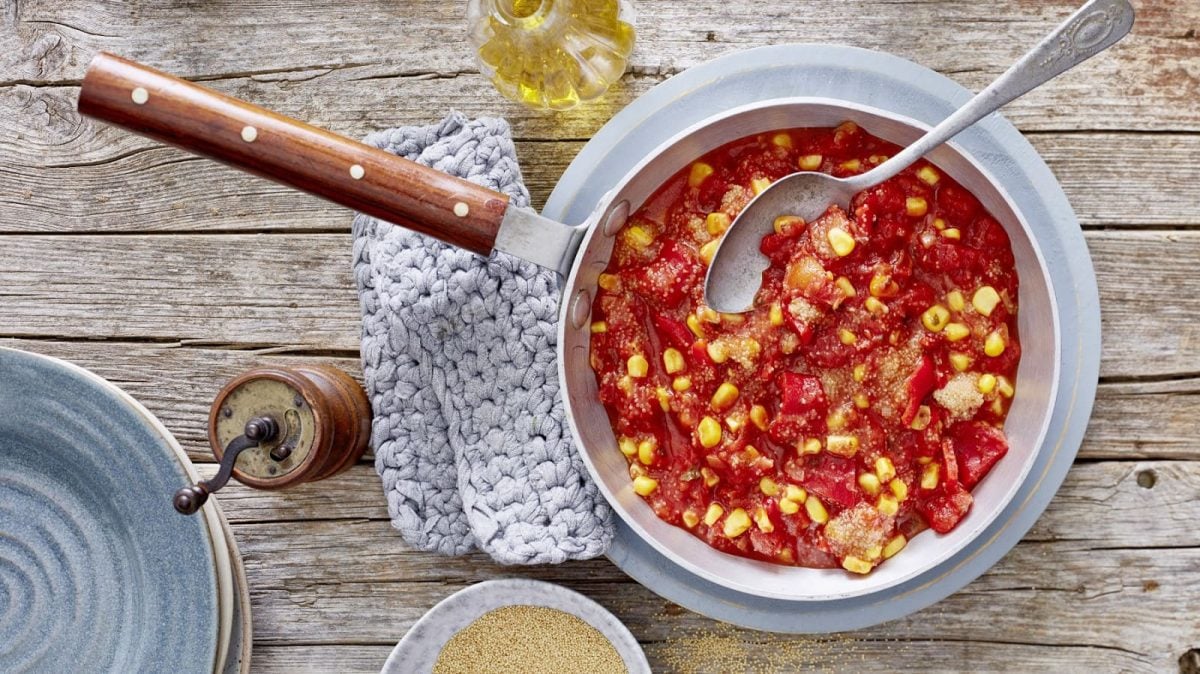 Eine Pfanne gefüllt mit Amaranth mit Paprika und Mais auf einem hölzernen Hintergrund. Links davon ein Topflappen, eine Pfeffermühle und Teller. Oben eine Flasche mit Öl und unten auf dem Bild eine kleine Schüssel mit Amaranth-Körnern.
