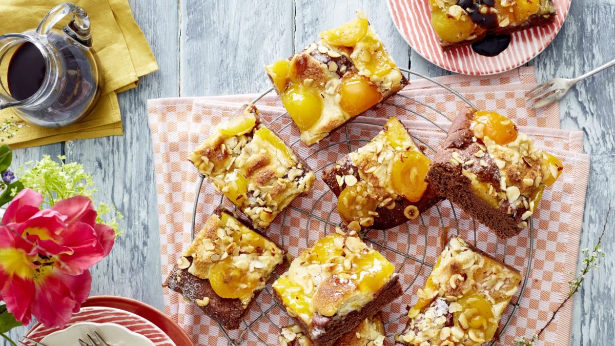 Acht Aprikosen-Schoko-Butterkuchen Stücke auf einem Kuchengitter auf einem orange/weißem Geschirrtuch. Links davon Blumen, eine Kaffeekanne und Teller. Rechts oben ein Teller mit einem Kuchenstück und einer Dessertgabel.