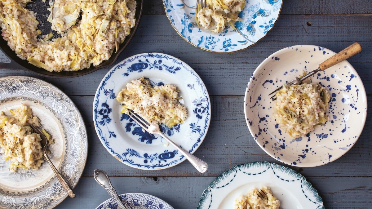 Mehrere blau-weiß-gemusterte Teller mit Artischocken-Risotto und Gabeln in der Draufsicht, außerdem eine große Pfanne mit mehr Risotto.