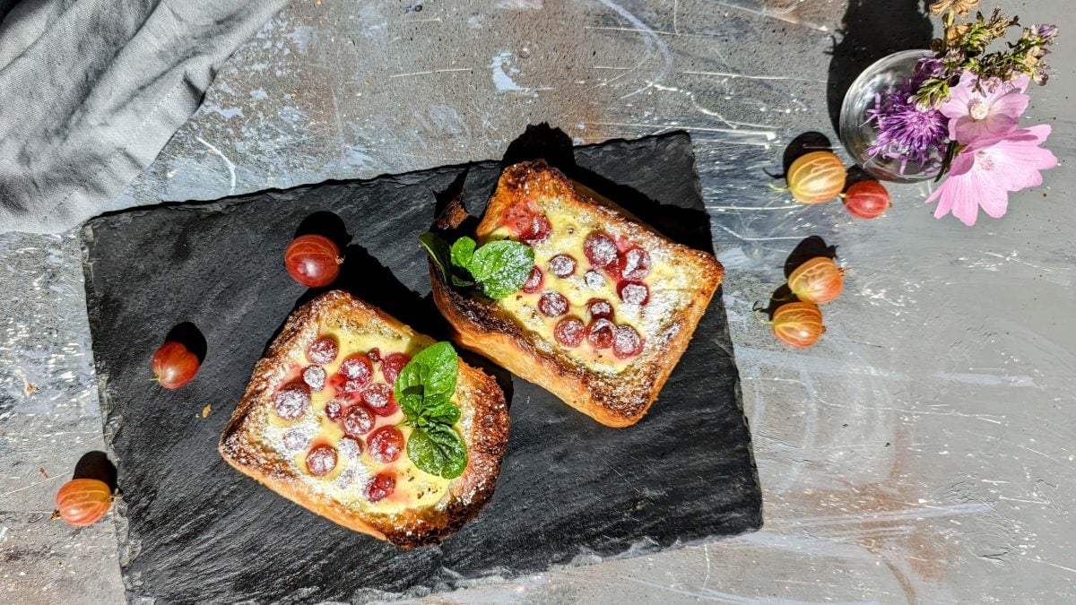 Zwei Brioche-Scheiben mit Quarkcreme und Stachelbeeren überbacken auf einer schwarzen Schieferplatte und grauem Untergrund von oben fotografiert.