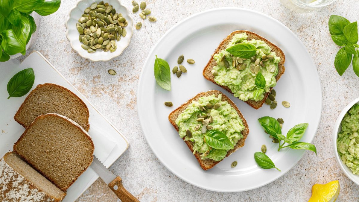 Zwei Brotscheiben mit Basilikum-Avocado, Kürbiskernen und Basilikumblättern auf einem Teller in der Draufsicht, drumherum ein angeschnittener Brotlaib, ein Stück Zitrone, eine Schüssel mit der Guacamole und Basilikum.