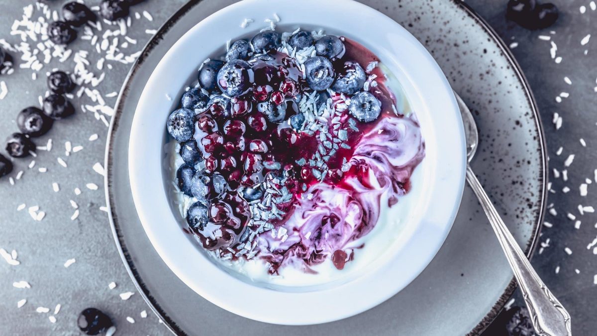 Draufsicht: eine weiße Schale Blaubeer-Kokos-Granola mit Quinoa steht auf einem hellem Teller, der auf dunklem Untergrund aus Holz steht.