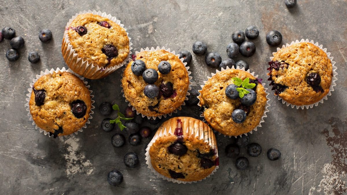Mehrere Blaubeermuffins auf einem grauen Steintisch mit frischen Blaubeeren und Minze.