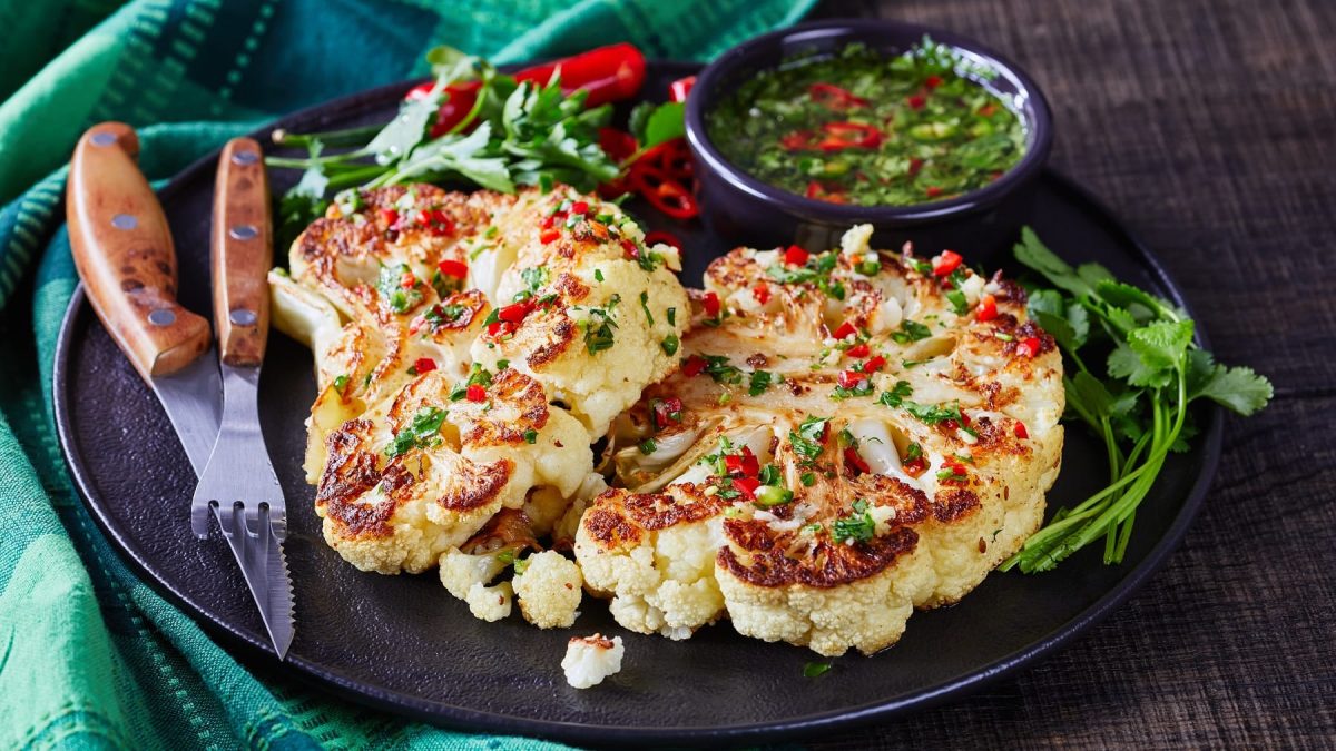 Zwei Blumenkohlsteaks auf einem schwarzen Teller mit asiatischem Dip und dunklem Untergrund. Daneben eine türkisfarbene Serviette.