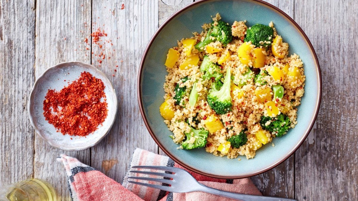 Eine Brokkoli-Bowl mit Couscous auf einem hölzernen Hintergrund. Links von der blauen Schüssel steht eine kleine Schüssel gefüllt mit Chiliflocken. Unten ein Geschirrtuch mit einer Gabel.