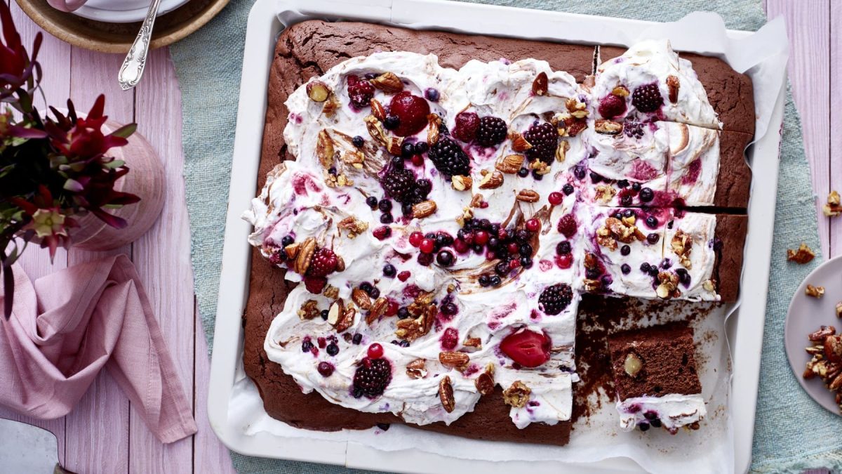Ein weißes Blech mit Brownies auf einem hellblauen Geschirrtuch auf einem rosa Untergrund. Links davon eine kleine Pflanze.