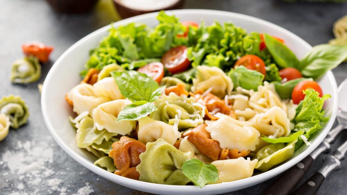 Ein weißer Teller mit dem Tortellini-Salat auf einem grauen Steintisch. Daneben eine Schale mit dem Dressing und einzelne Tortellini.