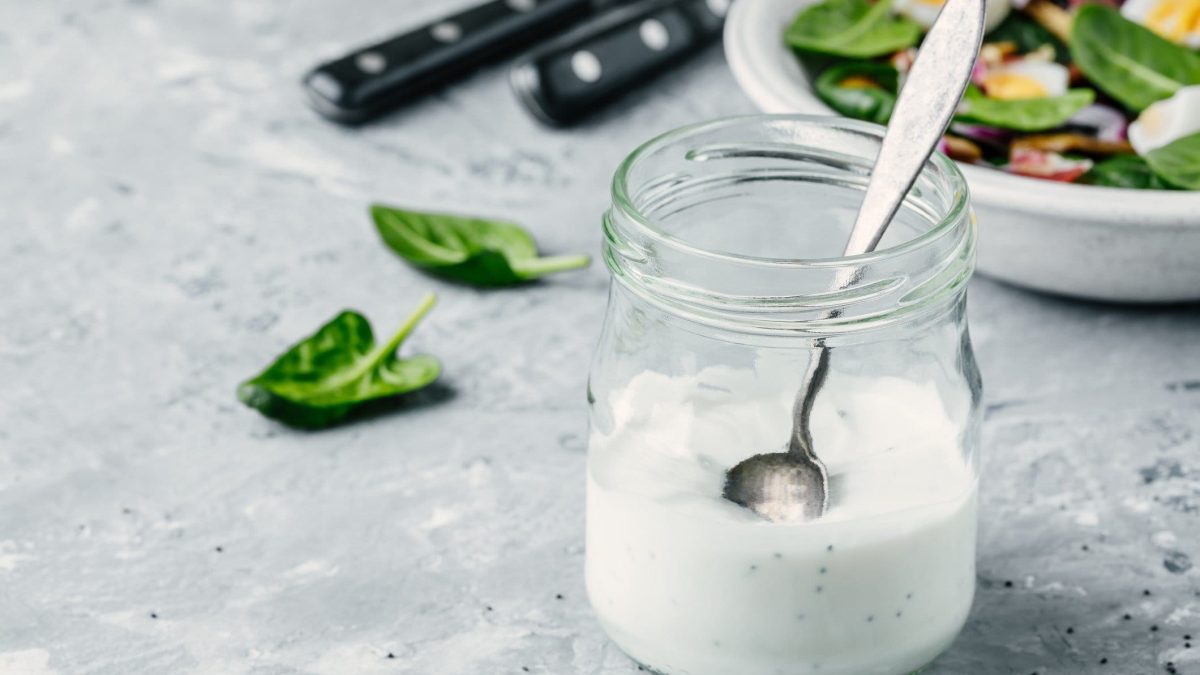 Buttermilch-Dressing mit Mohn in einem Glas mit Löffel. Das Glas steht auf hellem Untergrund, im Hintergrund sieht man eine Schale mit Salat.