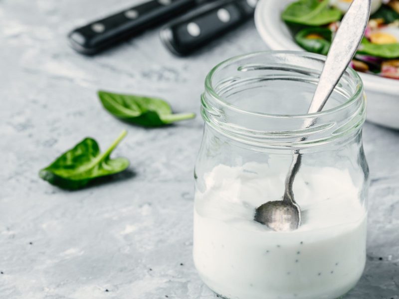 Buttermilch-Dressing mit Mohn in einem Glas mit Löffel. Das Glas steht auf hellem Untergrund, im Hintergrund sieht man eine Schale mit Salat.