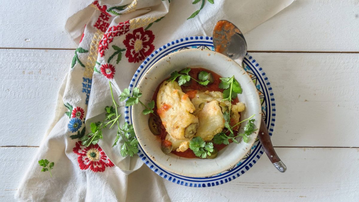 Chiles Rellenos con Queso, gefüllte Käsepaprika aus Mexiko, auf einem tiefen Teller, darin auch Tomatensauce, darauf ein Löffel und Koriander, daneben ein Geschirrtuch, alles Draufsicht.