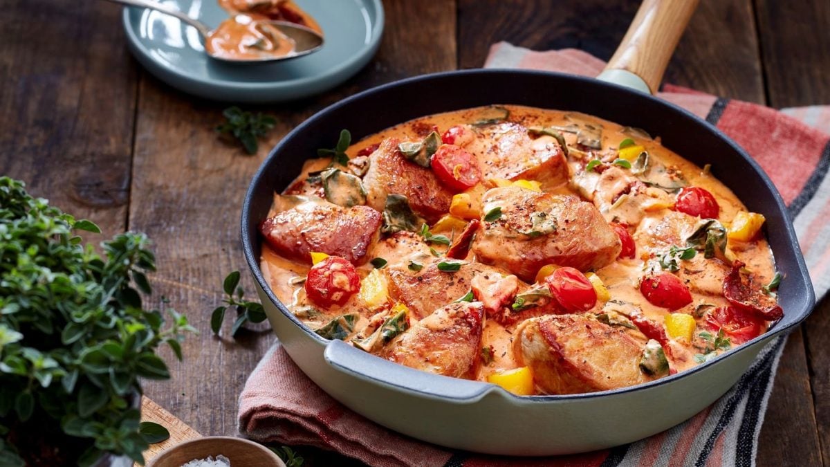 Eine schwarze Pfanne auf einem roten Geschirrtuch auf einem hölzernen Hintergrund. Die Pfanne ist gefüllt mit Pute in einer cremigen Tomatensauce. Links davon ein Kräuterstrauch.