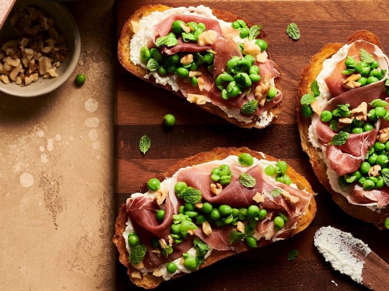 Ein Holzbrett mit Crostini mit Prosciutto und Erbsen auf einem Holztisch.