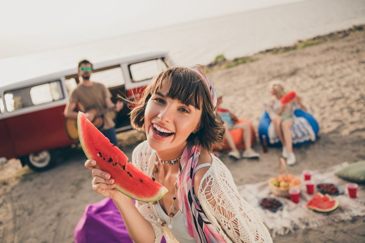 Frontal: Junge Menschen picknicken am Strand. Im Vordergrund ist eine junge Frau, die in eine Melonen-Scheibe in der Hand hält. Im Hintergrund ist ein VW-Bus.
