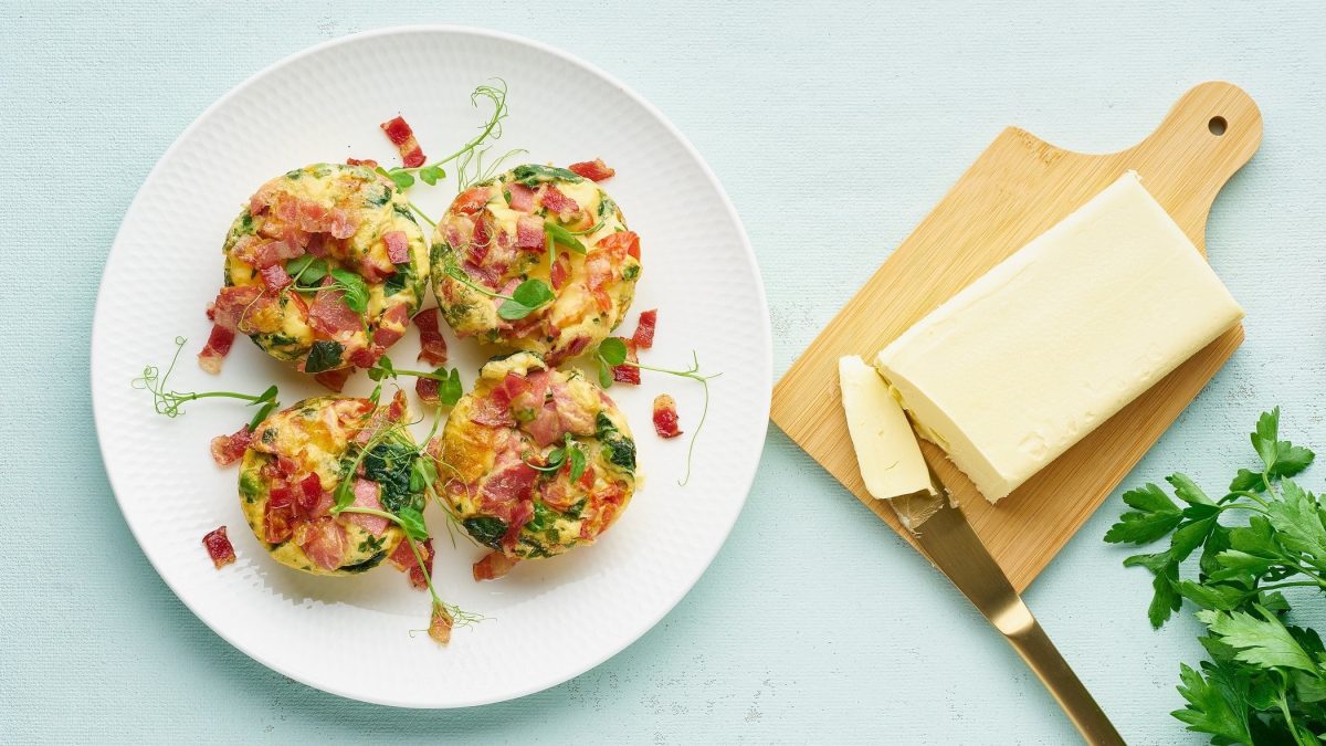 Draufsicht: Ein weißer Teller mit vier Eier-Muffins mit Speck und Spinat steht auf einem hellblauen Untergrund. Daneben liegen eine Butter auf einem Holzbrettchen, ein Küchenmesser und einige Blätter Petersilie.