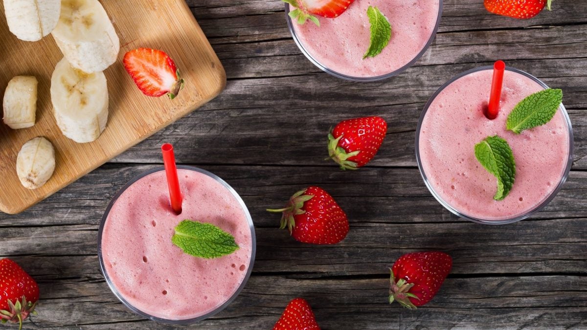 Mehrere Gläser mit dem Smoothie auf einem dunklen Holztisch mit Banane und Erdbeeren.