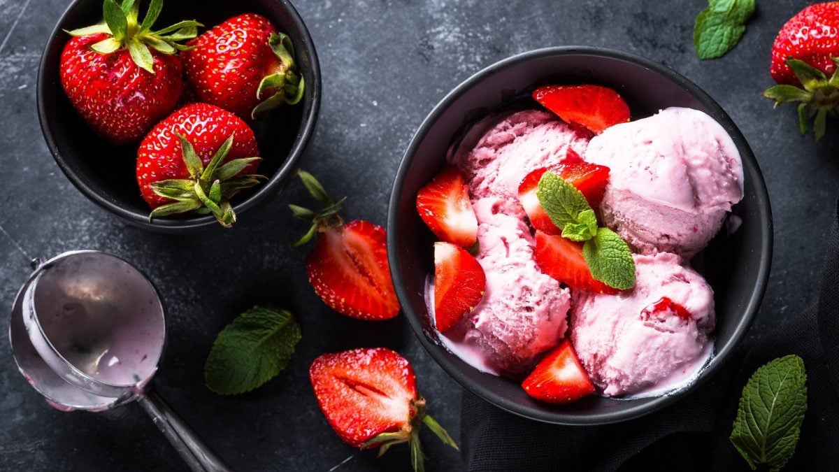 Eine Schüssel mit drei Kugeln Erdbeer-Nicecream und frischen Erdbeeren, daneben eine Schale mit Erdbeeren, ein Eisportionierer. Alles Draufsicht.