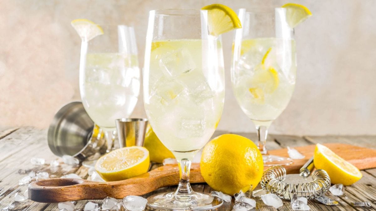 Drei French Spritz in Gläsern mit Ztronenscheiben vor hellem Hintergrund. Im Bild ganze Zitronen, geschnittene Zitronen und Barwerkzeuge. Frontalansicht.