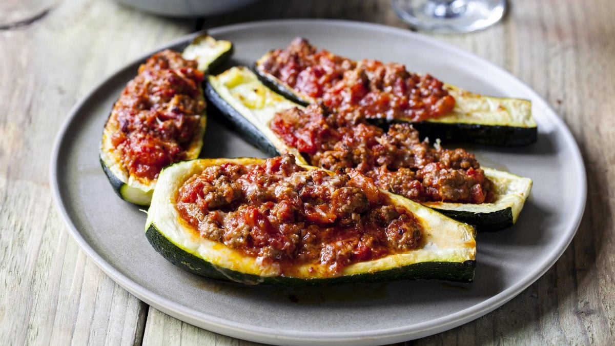 Frontalsicht: Auf einem Holztisch steht ein rustikaler Teller mit vier halben gefüllte Zucchini mit Lamm-Hackfleisch.