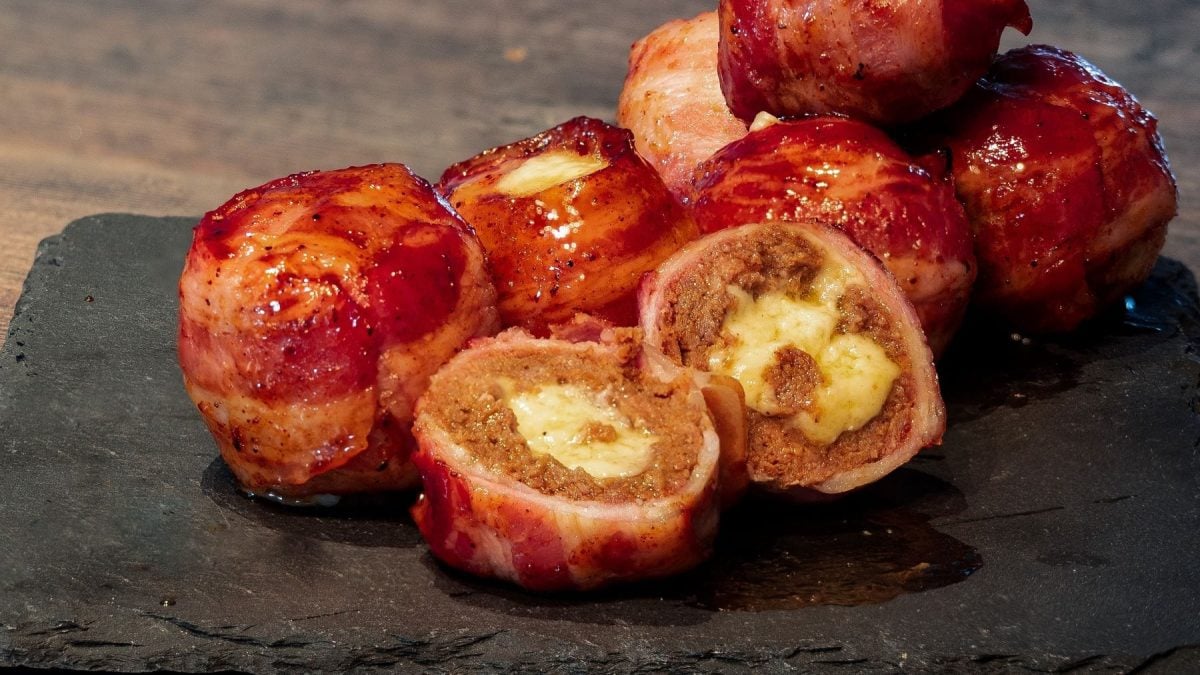 Gegrillte Frikadellen mit Käsefüllung und Zwiebel-Baconmantel auf Schieferplatte auf Holz. Eine Frikadelle aufgeschnitten dem Betrachter zugewandt. Frontalansicht.