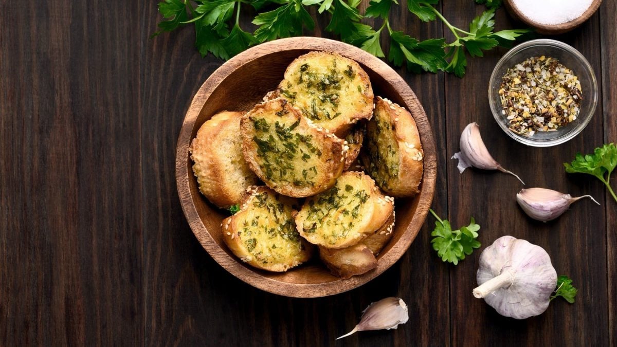 Draufsicht: Dunkler Untergrund, eine Korb mit geröstetem Knoblauchbrot mit Käse, frischer Knoblauch und frische Petersilie.