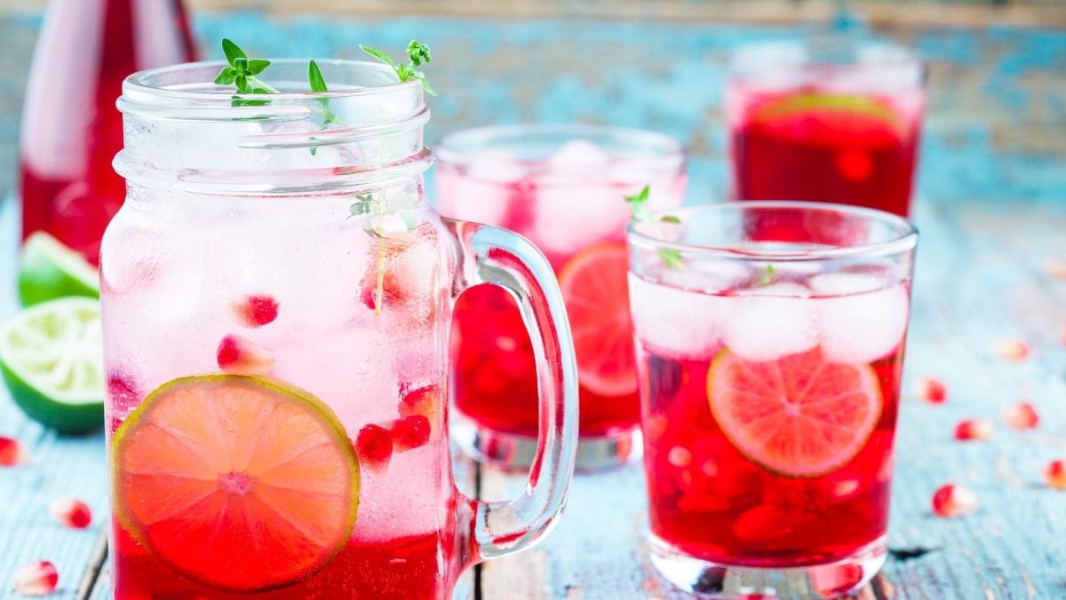 Frontal: Mehrer Gläse und ein Krug Granatapfel-Limetten-Drink stehen auf einem hellblauen Untergrund. Drum herum liegen Granatapfelkerne.