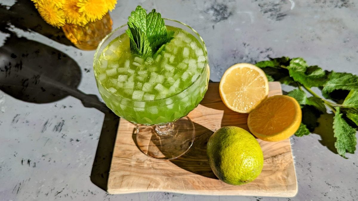 Der Green Limoni auf einem Holzbrett mit grauem Untergrund von schräg oben fotografiert. Im Hintergrund Zitronenmelisse und gelbe Blumen.