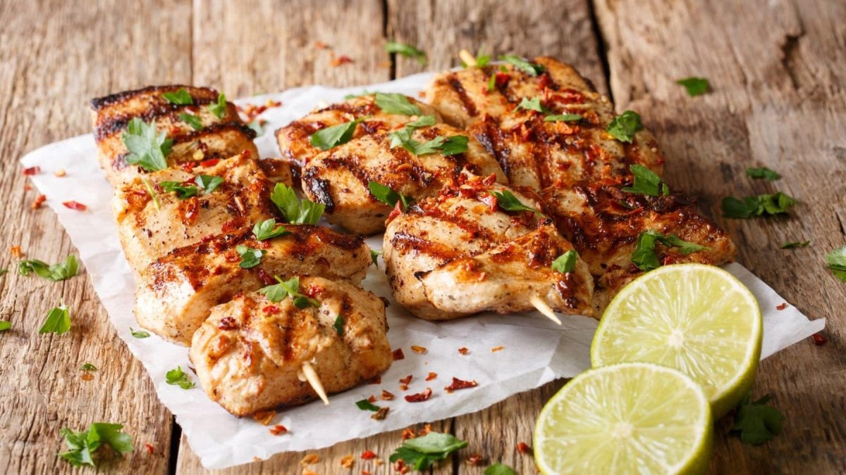 Zwei Hähnchenspieße in Joghurt-Kräuter-Marinade mit frischen Kräutern auf weißem Teller auf Holt. Im Bild eine halbierte Zitrone. Draufsicht.