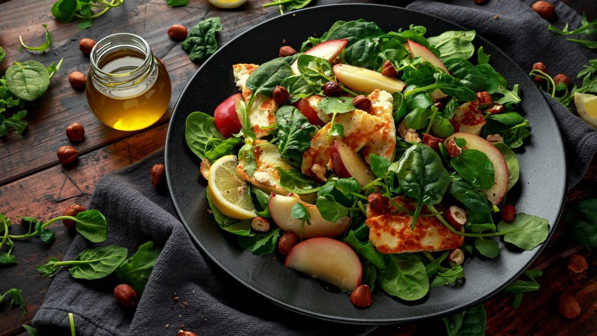 Ein schwarzer Teller mit dem Halloumi-Salat auf einem dunklen Holztisch mit Spinat und Haselnüssen.