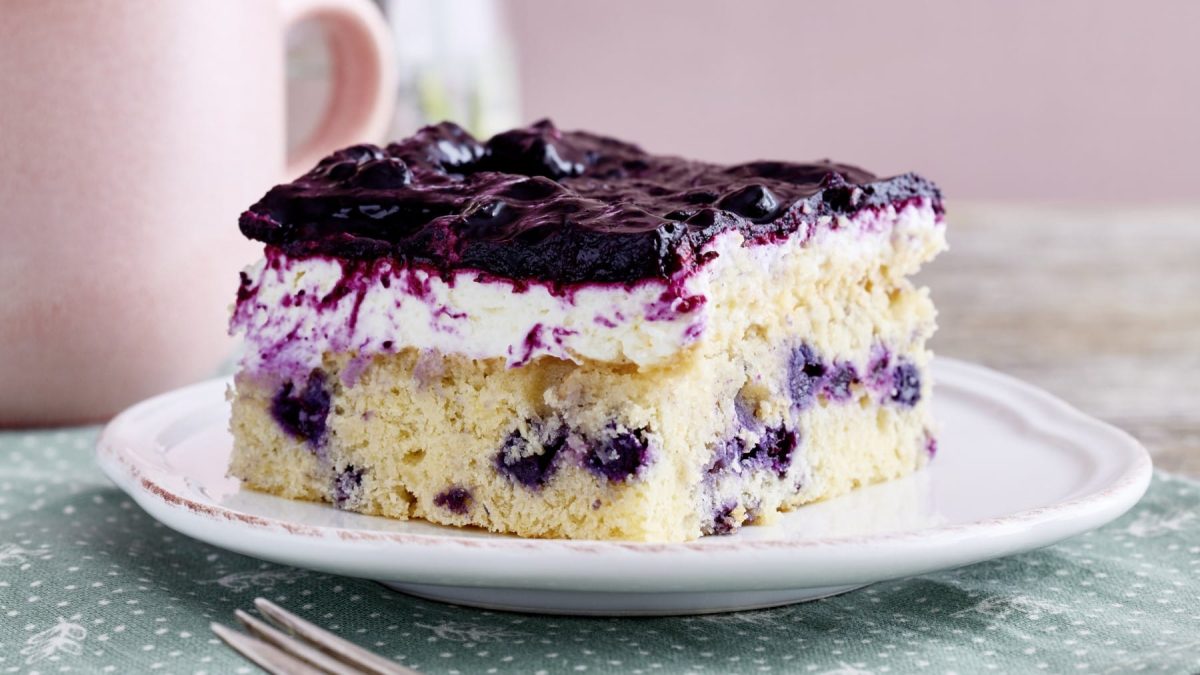 Ein Stück von einem Heidelbeer-Quarkkuchen auf einem weißen Teller von vorne fotografiert auf einer grünen Tischdecke mit weißen Punkten. Hinten links eine rosarote Tasse.