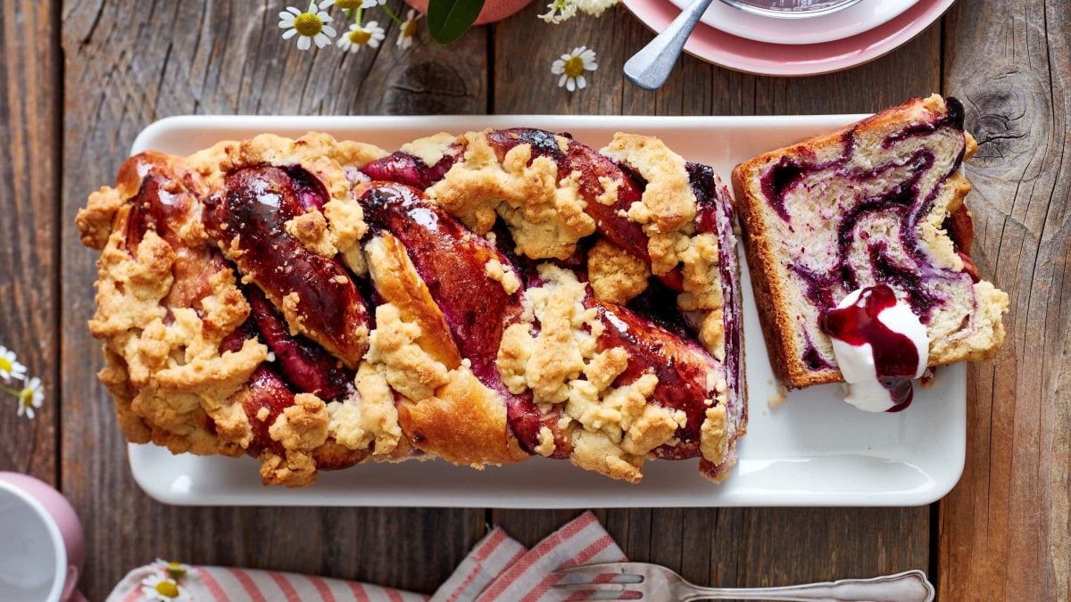 Ein Heidelbeer-Zopf mit Zitronen-Streusel auf einem weißen Servierteller auf einem hölzernen Hintergrund. Links unten eine Tasse und ein Geschirrtuch mit einer Dessertgabel. Oben auf dem Foto Untertassen und Gänseblümchen.