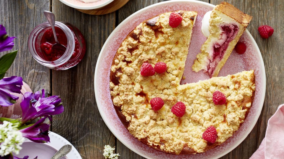 Ein Himbeer-Käsekuchen auf einem hellroten Servierteller auf einem hölzernen Hintergrund. Links davon ein Glas gefüllt mit Himbeerkompott und Blumen.
