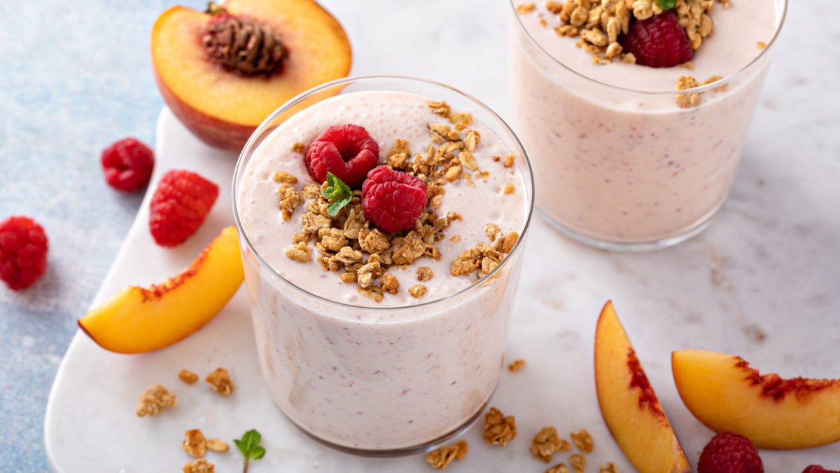 Draufsicht: Zwei Gläser Himbeer-Pfirsich-Smoothie. Daneben liegen Pfirsich-Spalten, Himbeeren und Granola.