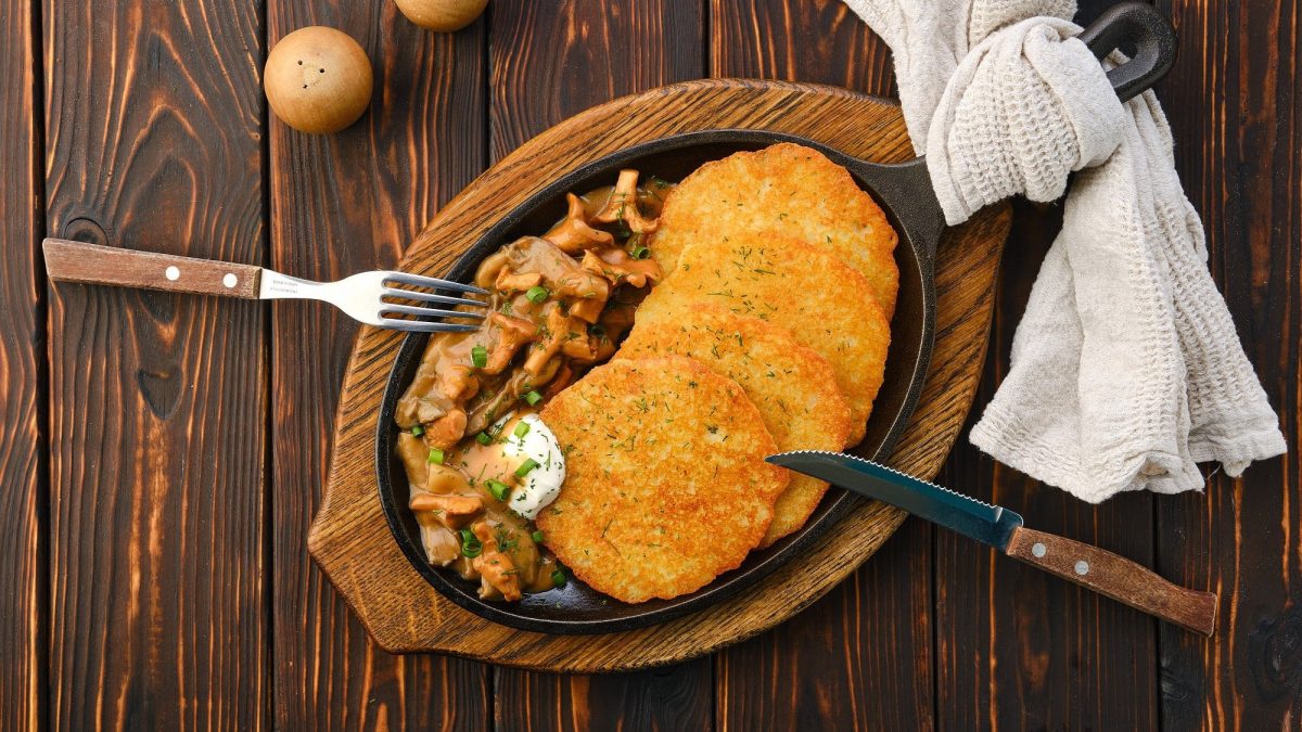 Eine gusseiserne Pfanne mit 3 Kartoffelpuffern mit Pfifferling-Rahmsauce, einem Klecks Schmand und Kräutern, daneben Messer und Gabel, Gewürzstreuer und ein Tuch, alles Draufsicht.
