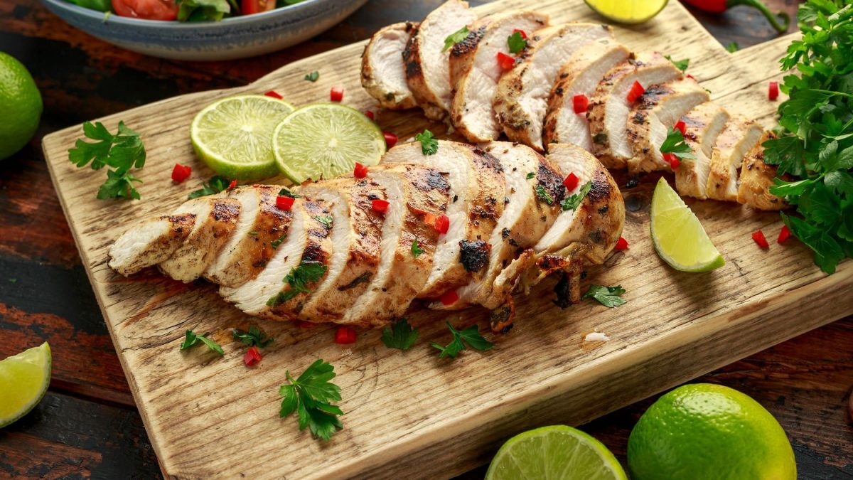 Zwei gegrillte und in Streifen geschnittene Hähnchenbrüste auf einem Holzschneidebrett mit Limetten und Kräutern.