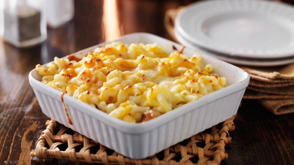 Makkaroni-Schinken-Käse-Auflauf einfach mal ohne Aufwand in weißer Auflaufform auf Holzuntersetzer und Tisch. Im Bild weiße Teller, Pfeffer- und Salzstreuer und Servietten. Frontalansicht.