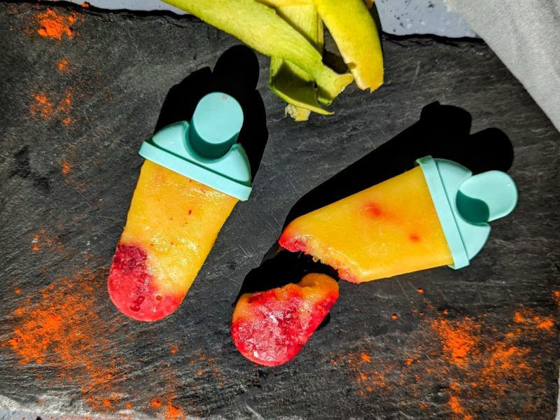 Zwei Mango-Eis mit Himbeere und Cayenne auf einer schwarzen Schieferplatte und grauem Untergrund. Daneben Mangoschalen und Cayennepfeffer.