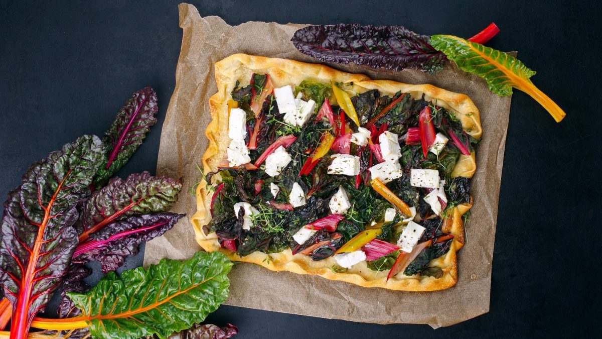 Ein Mangold-Flammkuchen mit Feta auf Backpapier in der Draufsicht, daneben frischer bunter Mangold.