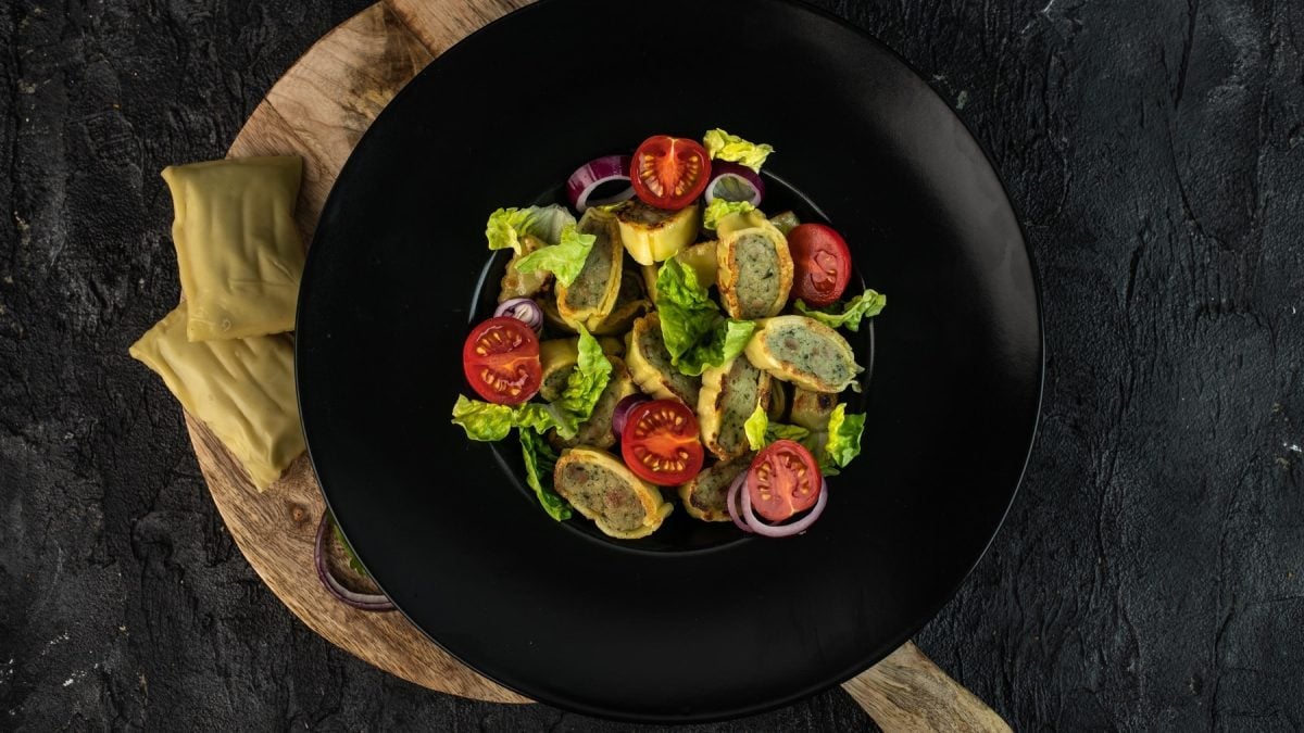 Ein schwarzer Teller mit dem Maultaschen-Salat auf einem Holzbrett und schwarzen Untergrund.