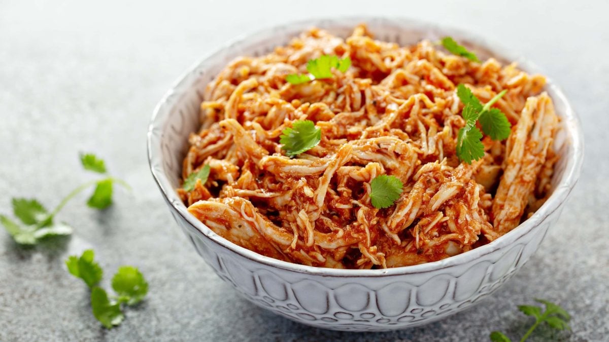 Eine weiße Schüssel mit dem Pulled Chicken auf einem grauen Tisch mit einigen Korianderstielen.