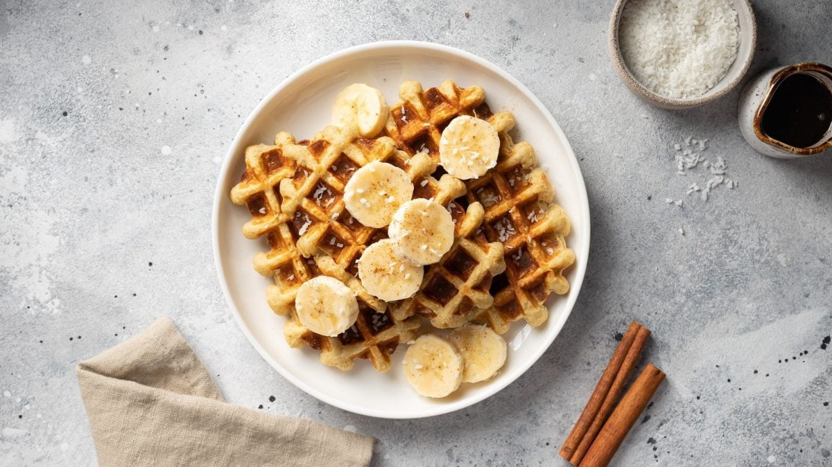 Milchreiswaffeln auf einem Teller mit Bananenscheiben und Kokos, daneben eine Schale Kokosraspeln, eine Serviette und Zimtstangen, alles Draufsicht.