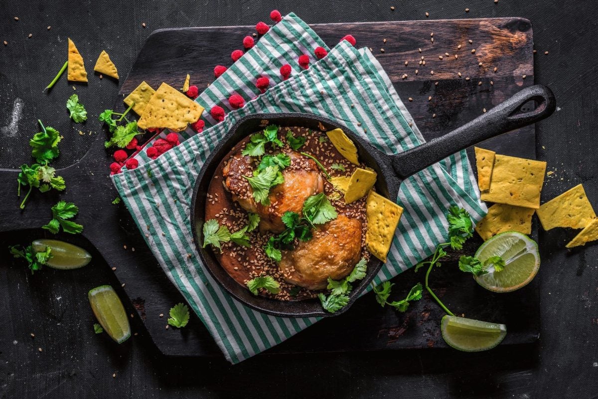 Mexikanisches mole poblano con Pollo, Hähnchenfleisch mit Schokoladensauce, in einer gusseisernen Pfanne, mit Nachos, Koriander und Sesam, Draufsicht.