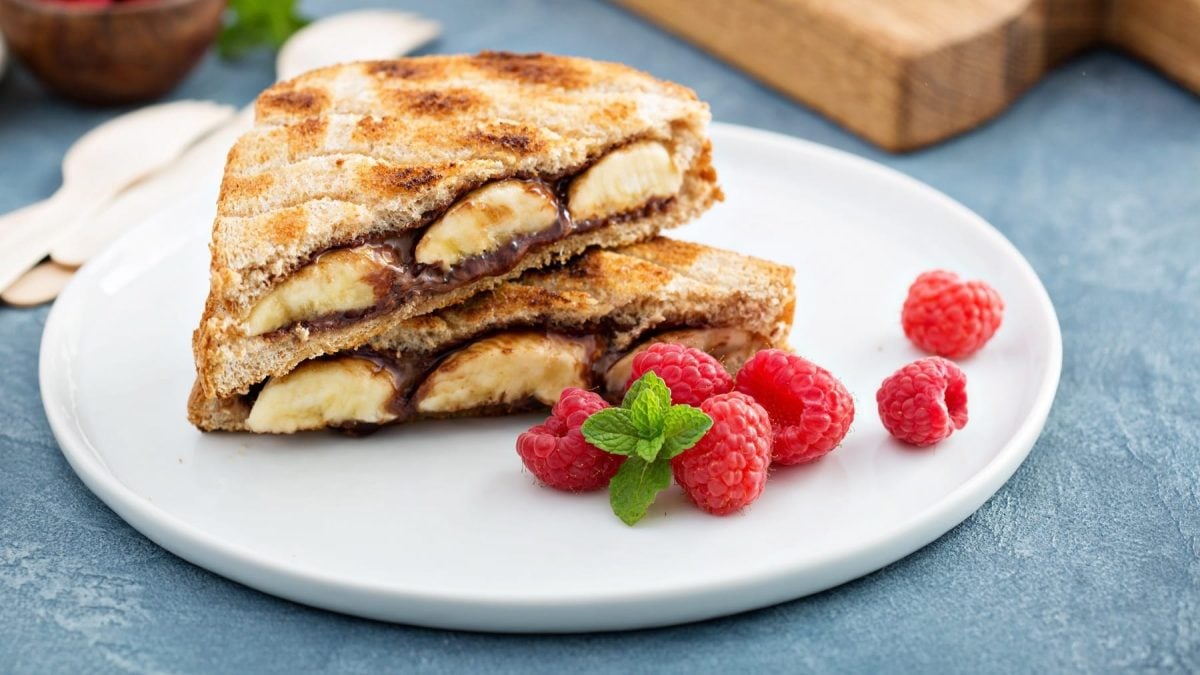 Ein angeschnittenes Bananen-Nutella-Toast auf einem weißen Teller mit frischen Himbeeren.