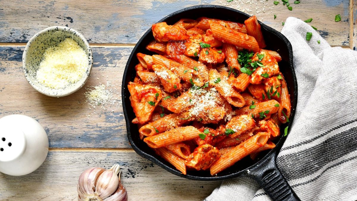 Eine schwarze gusseiserne Pfanne mit der One-Pot-Pasta auf einem grau-blauem Holztisch. Daneben eine Schale mit Parmesan sowie Salz und frischer Knoblauch.